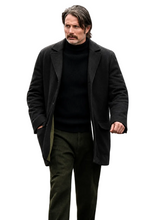 A man wearing a dark charcoal wool-blend coat over a black turtleneck sweater and dark olive trousers, standing against a solid black background.