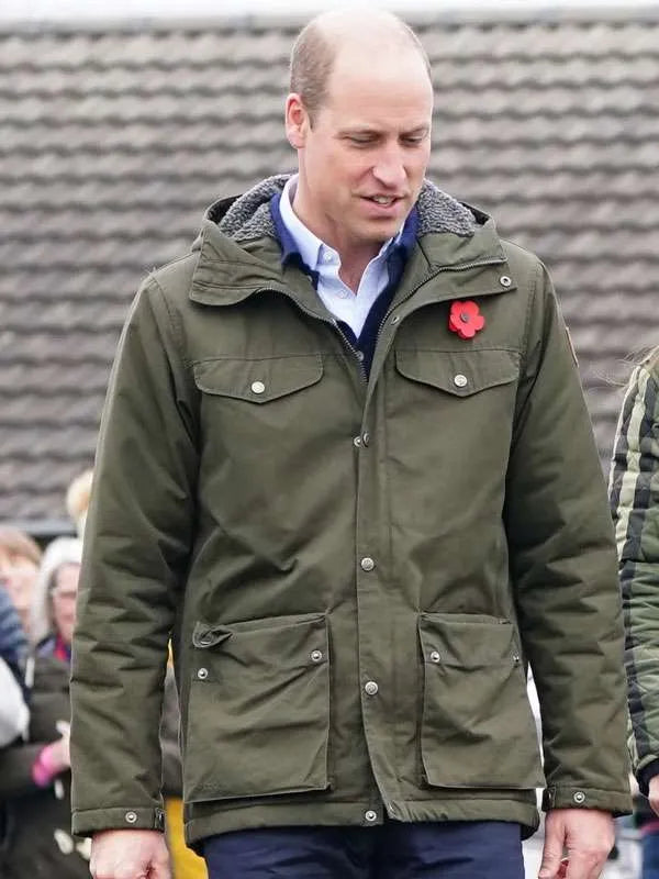 A man wearing an olive green hooded utility jacket with snap buttons, a blue shirt underneath, and a red poppy pin on the chest, standing outdoors in front of a gray tiled wall.