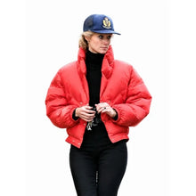 A woman wearing a bright red cropped puffer jacket over a black turtleneck and black pants, paired with a navy blue baseball cap, set against a plain white background.