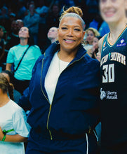 A woman smiling in a navy blue zip-up jacket with gold zippers, standing in an indoor sports arena. Another basketball player in a green New York jersey is partially visible on the right.