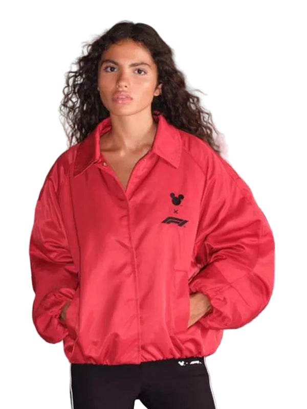 A woman wearing a bright red nylon jacket with a black embroidered logo on the left chest, paired with black track pants, standing in an industrial setting.
