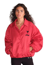 A woman wearing a bright red nylon jacket with a black embroidered logo on the left chest, paired with black track pants, standing in an industrial setting.