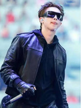 A man wearing a black leather jacket, black hoodie, black gloves, and black sunglasses, holding a microphone on a stage.