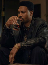 A man wearing a black leather jacket, black shirt, and gold jewelry sits and drinks from a glass of whiskey.