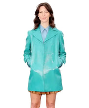 A woman stands outdoors wearing a bright turquoise-green raincoat over a light blue collared shirt and a brown pleated mini skirt. The background shows a blurred cityscape.