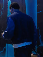 A man viewed from behind wearing a dark blue and black varsity-style jacket over a white shirt, standing in a dimly lit environment with blue lighting.