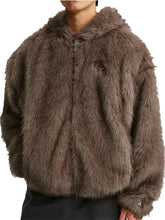 A person wearing a shaggy faux-fur cropped jacket in a taupe-brown color, paired with dark pants, against a light gray studio background.