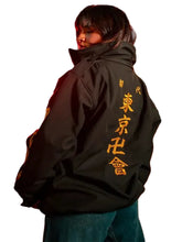 A person standing on a city street at night wearing an oversized black jacket with large gold Japanese characters printed down the back, paired with blue jeans.