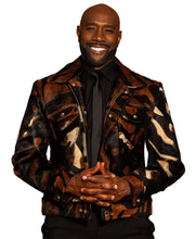A smiling man wearing a black, brown, and beige animal-print faux fur jacket over a black shirt and tie, standing against a solid brown studio background.