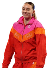 A woman wearing a bright red, orange, and pink zip-up athletic jacket with the number 26 on the front, standing on a stage-like setting.