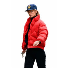 A woman wearing a bright red puffer jacket, black pants, and a navy blue baseball cap with a gold crest, isolated on a white background.