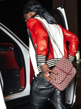 A man wearing a red and white leather jacket, black leather pants, and a black headwrap walks beside an open car door while carrying a pink Goyard shoulder bag with the brand's signature monogram pattern.