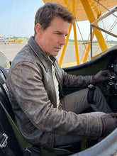 A man wearing a distressed brown leather jacket and gloves sits inside the cockpit of a vintage yellow biplane on an airfield.