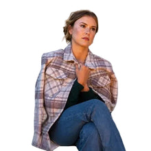A woman wearing a muted plaid button-up shirt in beige, brown, and gray tones, layered over a dark green top and blue jeans, sitting against a plain off-white wall.