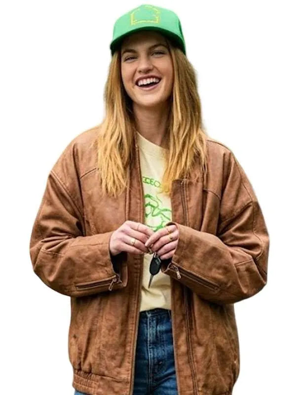 A woman wearing an oversized brown faux-leather jacket, a green baseball cap, a graphic t-shirt, and blue jeans, smiling against a solid white background.