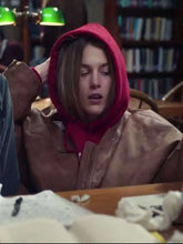 A person wearing a brown leather jacket and a bright pink hoodie sits at a wooden table in a library or study, with books visible in the background.