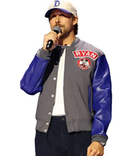 A man on stage wearing a gray and blue varsity-style jacket with 'RYAN' and a cartoon character patch on the chest, a white t-shirt, dark pants, and a white cap with a blue 'D' logo, holding a microphone.