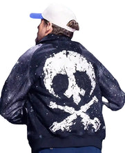 A man shown from behind wearing a black bomber jacket with a large distressed white skull and crossbones graphic on the back, paired with a white baseball cap.