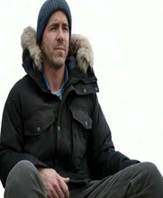 A man wearing a dark winter parka with a fur-trimmed hood, a blue knit beanie, and dark pants, seated against a plain light background.