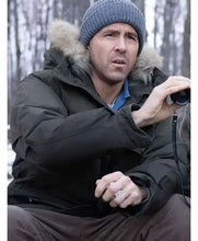 A man wearing a dark winter parka with a fur-trimmed hood, a blue knit beanie, and brown pants, holding binoculars in an outdoor snowy forest setting.