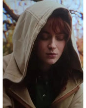 A person wearing a beige hooded jacket with the hood up, photographed outdoors with blurred autumn trees in the background.