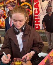 A woman in a brown jacket and white t-shirt signs a book at an event, with movie posters in the background.