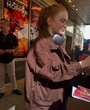 A woman with red hair in a ponytail wearing a dusty rose-brown jacket and a silver headphones around her neck, standing in front of a movie poster for 'The Book of Mormon'.