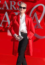 A woman stands on a red carpet wearing a bright red coat over a white blouse and black pants, with large metallic letters behind her.