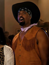 A man wearing a brown suede fringe vest, a black cowboy hat, a black bandana, and a light pink bandana around his neck, captured in a warm indoor setting.