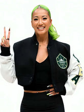 A smiling woman with bright green hair poses in a black and white varsity-style bomber jacket with a green shield logo on the chest, against a plain white background.