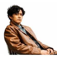 A man with dark curly hair wearing a brown leather jacket over a blue-gray button-down shirt, posed against a solid white background.