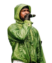 A man wearing a shiny green hooded rain jacket and black gloves holds a microphone while performing in the rain.
