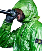 A person wearing a shiny bright green hooded rain jacket and black gloves, holding a microphone close to their mouth against a dark studio background. A Stone Island logo patch is visible on the sleeve.