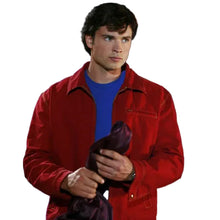 A man wearing a red corduroy utility jacket over a blue t-shirt, holding a dark purple garment against a dark blue curtain background with a red balloon visible in the upper right.