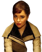 A close-up portrait of a woman with short brown hair wearing a beige jacket with a dark fuzzy collar, set against a dark background.