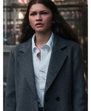 A woman wearing a dark gray double-breasted coat over a white button-down shirt, standing outdoors with a blurred urban background.