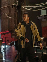 A man standing on a city street at night wearing a tan and black bomber jacket over a black shirt and dark pants.