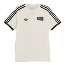 An off-white short-sleeve t-shirt with black three-stripes on the sleeves, a black Adidas trefoil logo on the left chest, and a black 'oasis' patch on the right chest, shown against a light beige background.