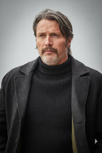 A man wearing a dark charcoal gray wool-blend coat over a black ribbed turtleneck sweater, standing outdoors.