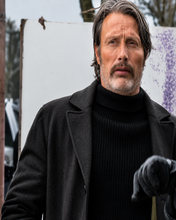 A man wearing a dark charcoal gray wool coat over a black ribbed turtleneck sweater, with a black glove on one hand, photographed outdoors against a white sign and trees.
