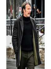 A man wearing a dark charcoal gray wool-blend coat over a black ribbed turtleneck sweater and olive green trousers, standing outdoors on a city street with snow patches and a railing in the background.