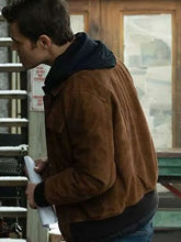 A man wearing a brown suede jacket over a black hoodie, standing in an outdoor or industrial setting.