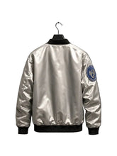 A shiny silver bomber jacket displayed on a black hanger against a dark background, with a blue and white circular patch on the left sleeve.