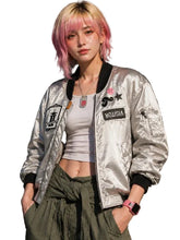 A model with pink-tinted hair wearing a shiny silver bomber jacket with black trim and various patches, paired with an olive green shirt tied at the waist and a white crop top, standing against a dark textured wall.