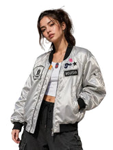 A woman wearing a shiny silver bomber jacket with black trim and multiple embroidered patches, standing in front of a graffiti-covered metal shutter.