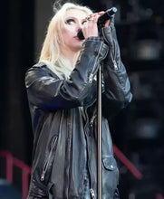 A blonde woman wearing a black leather jacket stands at a microphone on a stage, with a dark, blurred background.