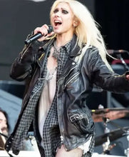 A blonde woman performing on stage, wearing a black leather motorcycle jacket over a plaid shirt and light-colored top, holding a microphone.