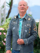 An older man wearing a gray embroidered western-style shirt jacket over a turquoise paisley button-up shirt, standing outdoors with blurred greenery and mountains in the background.