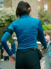 A woman seen from behind wearing a bright blue long-sleeve athletic top and black pants, with a black armband device on her left arm. A child in a gray uniform stands beside her in an outdoor street setting.