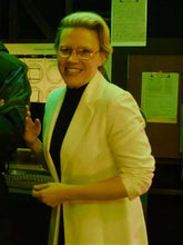 A woman with blonde hair and glasses, wearing a white blazer over a black top, smiling in what appears to be an office or laboratory setting.